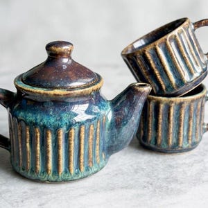 May include: A ceramic tea set with a teal, brown, and gold glaze. The set includes a teapot with a lid and two stacked mugs. The teapot and mugs have vertical ribbed detailing.