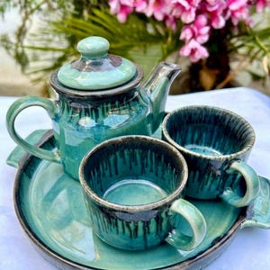 Puede incluir: Un juego de té de cerámica que consta de una tetera con tapa, dos tazas a juego y una bandeja. El conjunto presenta un esmalte brillante en tonos azul verdoso y turquesa con patrones de goteo de color marrón oscuro en los bordes.