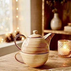 Puede incluir: Un juego de té de cerámica para una persona que consiste en una tetera apilada directamente sobre una taza a juego. Ambas piezas tienen un acabado en esmalte color crema con rayas horizontales de pinceladas marrones y un asa redondeada.