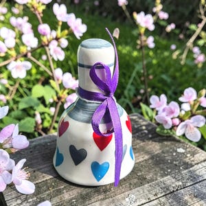 Puede incluir: Adorno de cerámica en forma de campana con base blanca, adornado con diseños de corazones rojos, negros y azules. La parte superior es blanca con rayas grises, y una cinta morada está atada alrededor del cuello. La campana está sobre una superficie de madera.