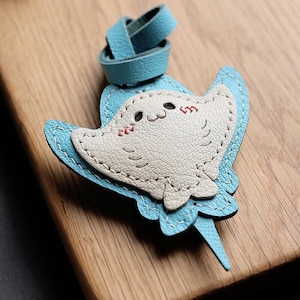 May include: A handmade leather stingray keychain in light blue and off-white. The stingray has a smiling face with pink cheeks and is stitched with white thread. The keychain has a loop of light blue leather.