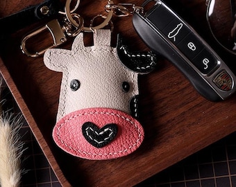 Pode incluir: Um porta-chaves de couro em forma de vaca com um nariz rosa em forma de coração e detalhes em preto. O porta-chaves está preso a um anel dourado e a uma alça de couro preta. Uma chave do carro e óculos também estão na imagem.