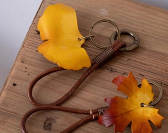 May include: Two leather leaf keychains. One is yellow, resembling a ginkgo leaf, and the other is a maple leaf with orange and yellow hues. Both have brown leather straps and metal key rings. The keychains rest on a wooden surface.