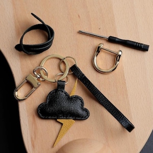 May include: A collection of accessories including a black leather cloud keychain with a yellow lightning bolt, a gold-colored horseshoe-shaped clasp, a black leather strap, a small screwdriver, and a coiled black leather cord. The items are arranged on a light wooden surface.