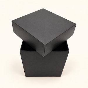 May include: A square, matte black gift box with a removable lid. The lid is positioned at an angle, partially resting on the open base of the box. The structure is made of solid black cardboard with clean, sharp edges and a smooth finish.