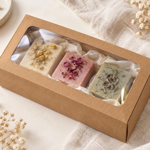 May include: A rectangular brown cardboard gift box with a clear plastic window containing three square bars of soap. The soaps are cream-colored with dried chamomile flowers, pink with dried rose petals, and sage green with dried lavender.