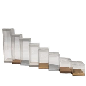 Puede incluir: Un conjunto de cajas de exhibición rectangulares transparentes de diferentes alturas. Cada caja tiene un cuerpo transparente y una base en tonos dorados, plateados y marrones. Las cajas están dispuestas en forma de escalera descendente.