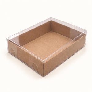 May include: A rectangular box with brown cardboard sides and a clear, transparent plastic lid. The base is constructed from light brown paperboard, featuring small semi-circular cutouts along the lower edges of the side panels.