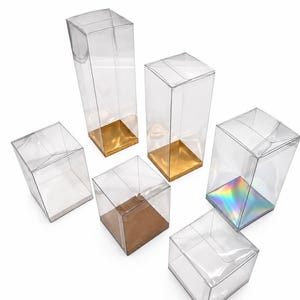 May include: A collection of six clear plastic display boxes of varying heights and widths. Two boxes feature gold bases, one has a metallic copper base, one has a holographic iridescent base, and two have plain white bases.