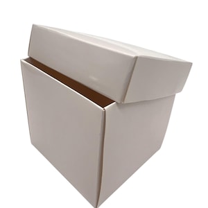 Peut inclure: Une boîte rectangulaire blanche avec un couvercle légèrement entrouvert. La boîte est faite d'un matériau lisse et mat et a un design simple et épuré. Le couvercle est incliné, révélant l'intérieur de la boîte.