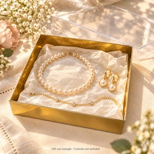 Puede incluir: Una caja de regalo dorada que contiene un collar de perlas, un par de pendientes colgantes dorados con detalles de perlas y una delicada pulsera de cadena dorada con pequeñas piedras transparentes de corte redondo engastadas.