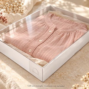May include: A light pink button-down shirt with a mandarin collar and textured fabric, neatly folded inside a rectangular white gift box lined with tissue paper. The shirt features three visible white buttons along the front placket.