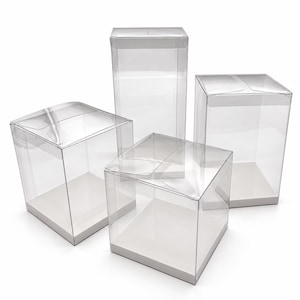 May include: A set of four clear plastic display boxes with white bases. The collection includes three square-shaped boxes of varying heights and one taller rectangular box, all featuring foldable top flaps for secure closure.