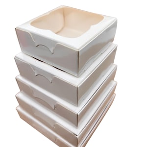 Op de afbeelding: Een stapel van vier witte kartonnen dozen met een glanzende afwerking. De bovenste doos heeft een helder venster. Elke doos heeft een gebogen uitsparing aan de zijkant voor gemakkelijk openen. De dozen zijn leeg en klaar voor gebruik.