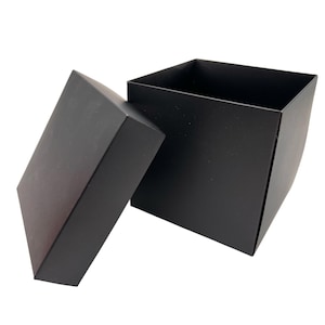 Peut inclure: Une boîte carrée noire avec le couvercle légèrement entrouvert, révélant l'intérieur. La boîte est faite d'un matériau mat et repose sur un fond blanc. Le couvercle est incliné, montrant la profondeur et la forme de la boîte.