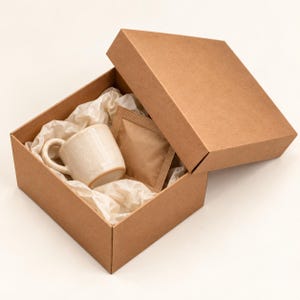 May include: A square brown cardboard gift box with a matching lid, containing a small, speckled off-white ceramic mug and a square brown paper packet nestled inside white crinkled tissue paper padding.