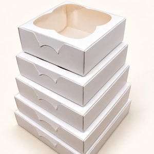 May include: A stack of five square white cardboard boxes, each featuring a clear plastic window on the top lid with a scalloped edge design. The boxes are uniform in size and shape, stacked vertically to create a single column of packaging.