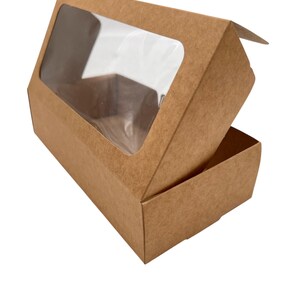 May include: A rectangular brown cardboard box with a clear plastic window on the top lid. The box is partially open, revealing the interior space. The exterior surface has a textured, natural paper finish in a light brown shade throughout.