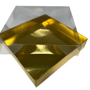 Puede incluir: Una tapa de plástico transparente descansa sobre una caja cuadrada dorada. La caja tiene un acabado brillante y metálico. La caja y la tapa están en ángulo, mostrando su forma geométrica. La caja es probablemente para embalaje o regalo.