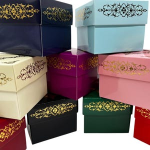 Peut inclure: Plusieurs boîtes cadeaux colorées, dans des tons de bleu marine, bleu clair, rose, rouge et noir. Chaque boîte est ornée d'un motif floral doré sur le couvercle. Les boîtes sont empilées, mettant en valeur leurs couleurs et motifs.