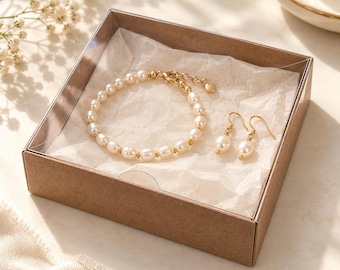 May include: A jewelry set consisting of a pearl bracelet and a pair of matching pearl drop earrings, presented in a square brown cardboard gift box with a clear lid. The bracelet features white pearls with gold-tone spacers.
