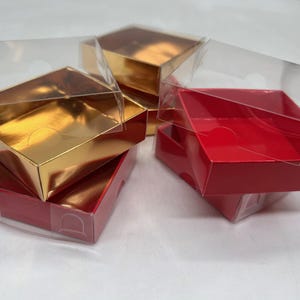 May include: A collection of small, square boxes with transparent plastic lids. Some are red, others gold. The boxes are stacked and slightly open, revealing their interiors.