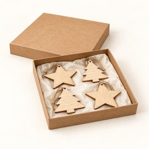 May include: A set of four light-colored wooden ornaments in a square cardboard box. The ornaments include two five-pointed stars and two stylized pine trees, each featuring a small hole at the top with a thin brown twine loop for hanging.