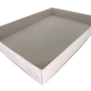 May include: A rectangular, white cardboard tray with a shallow depth. The tray has a smooth, reflective surface and small, curved cutouts on the sides for easy handling. The interior is a light, neutral color.