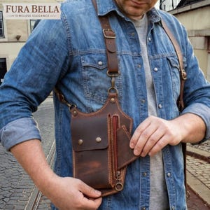 May include: A brown leather holster bag with adjustable straps, featuring a main compartment with a flap closure secured by two metal snaps, a zippered side pocket, and an additional small open pocket, worn over a blue denim shirt.