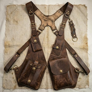 Op de afbeelding: Een schouderholsterharnas van bruin leer met twee hoofdvakken met drukknoopsluitingen, verstelbare riemen met metalen gespen en messingkleurige hardware. Het ontwerp bevat een centraal rugpaneel en extra kleine vakken voor organisatie.