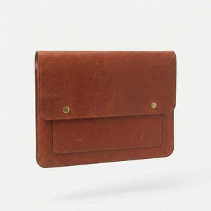May include: A brown leather laptop sleeve with a flap closure and a small pocket on the front. The sleeve is secured with two brass-colored snap closures. The leather has a natural, textured appearance.