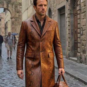 May include: A long, copper-brown coat with a textured snakeskin pattern, featuring a notched lapel, two front buttons, and two flap pockets. The coat is worn over a dark shirt and is paired with a brown leather duffel bag with a silver zipper.