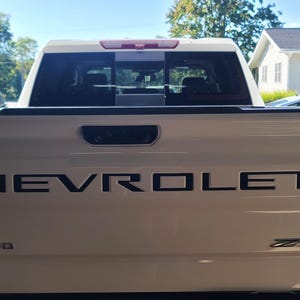 Könnte beinhalten: Rückansicht eines weißen Chevrolet Silverado-Trucks. Das Wort "CHEVROLET" ist in schwarzen Buchstaben auf der Heckklappe zu sehen. Das Wort "SILVERADO" steht darunter. Rote Rücklichter sind sichtbar.