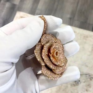 May include: A rose-shaped pendant with a sparkling, textured surface. The pendant is a warm gold color and is covered in small, glittering stones. The rose has multiple layers of petals and a detailed center. The pendant is held by a gloved hand.