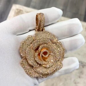 May include: A rose-shaped pendant with a bail, covered in small, sparkling stones. The pendant is a warm gold color, with a detailed design of rose petals. The bail is also covered in the same sparkling stones.