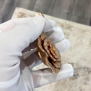 May include: A rose-shaped pendant with a gold finish, encrusted with small, sparkling gemstones. The pendant's design features layered petals, creating a three-dimensional effect. The jewelry is held by a gloved hand, showcasing the intricate details and craftsmanship.