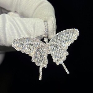 May include: A silver butterfly pendant with a sparkling, gem-studded design. The pendant features a detailed butterfly shape with intricate wing patterns. The gems are set closely together, creating a brilliant, eye-catching effect. The pendant is held against a dark background.