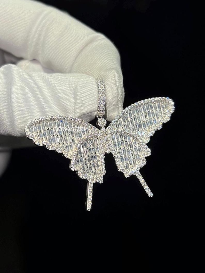 May include: A silver butterfly pendant with a sparkling, gem-studded design. The pendant features a detailed butterfly shape with intricate wing patterns. The gems are set closely together, creating a brilliant, eye-catching effect. The pendant is held against a dark background.