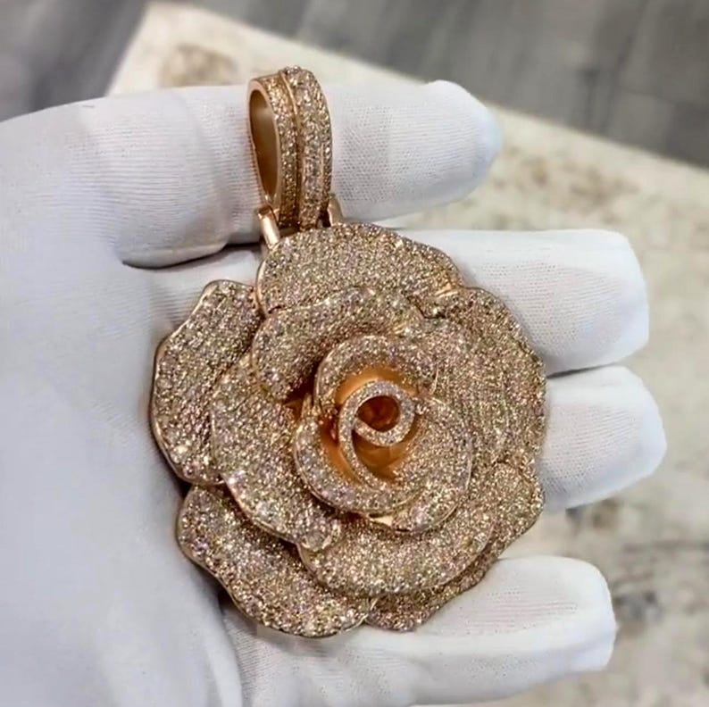 May include: A rose-shaped pendant with a gold-colored base and encrusted with numerous small, sparkling stones. The pendant is held by a gloved hand, showcasing the intricate details of the jewelry. The bail is also adorned with stones.