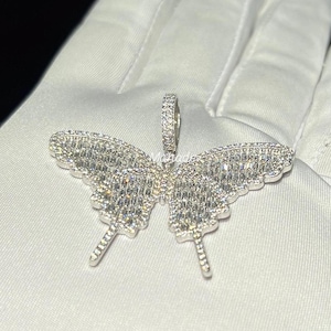 May include: A silver butterfly pendant with a sparkling, gem-like appearance. The pendant is intricately designed with a detailed wing pattern and a small loop for a chain. The butterfly pendant is displayed on a white surface.