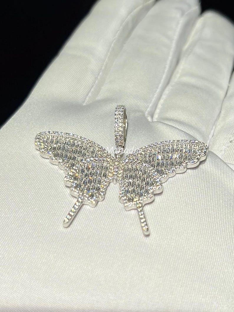 May include: A silver butterfly pendant with a sparkling, gem-like appearance. The pendant is intricately designed with a detailed wing pattern and a small loop for a chain. The butterfly pendant is displayed on a white surface.