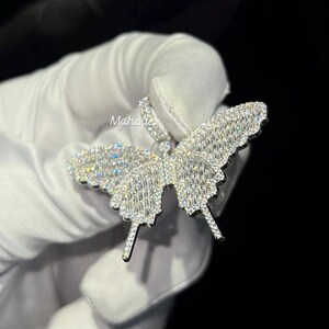 May include: A sparkling butterfly pendant, encrusted with numerous small, clear gemstones. The pendant's wings are spread wide, showcasing the intricate design and reflecting light. The jewelry is held against a dark background.