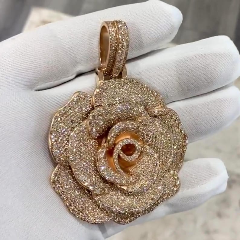 May include: A rose-shaped pendant with a rose gold finish, encrusted with numerous small, sparkling gemstones. The pendant is held by a gloved hand, showcasing the intricate details of the floral design and the brilliance of the gemstones.