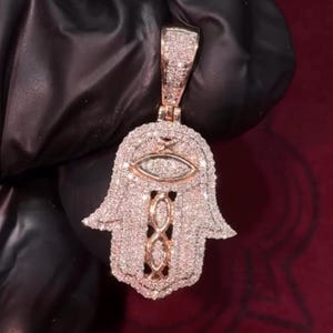 May include: A rose gold and silver pendant in the shape of a Hamsa hand, encrusted with small, sparkling stones. The pendant features an eye design in the center and an infinity symbol at the base. The background is a dark, textured surface.