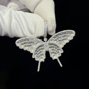 May include: A sparkling butterfly pendant with a silver finish. The wings are encrusted with numerous small, clear crystals, creating a dazzling effect. The pendant is held by a white gloved hand against a black background.
