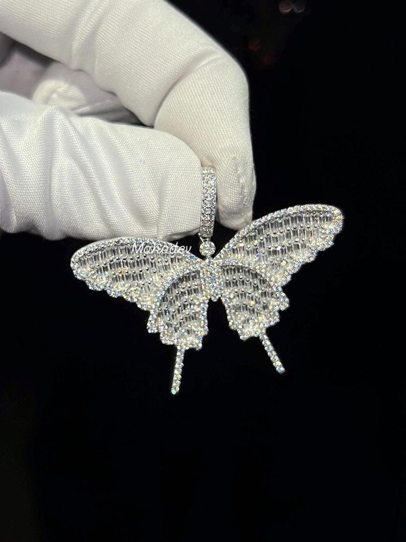 May include: A sparkling butterfly pendant with a silver finish. The wings are encrusted with numerous small, clear crystals, creating a dazzling effect. The pendant is held by a white gloved hand against a black background.