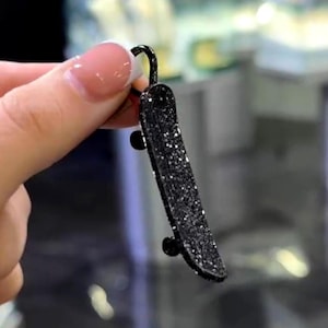 Könnte beinhalten: Ein Miniatur-Skateboard-Schlüsselanhänger mit schwarzem Glitzer-Finish. Das kleine Skateboard hat schwarze Räder und eine Schlaufe zum Befestigen an Schlüsseln oder einer Tasche. Der Glitzer verleiht dem Accessoire Glanz.