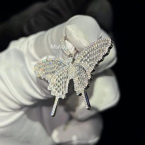 May include: A sparkling butterfly pendant, encrusted with numerous clear gemstones. The pendant is held against a dark background, showcasing its intricate design and reflective qualities. The butterfly's wings and body are detailed with a dazzling array of stones.