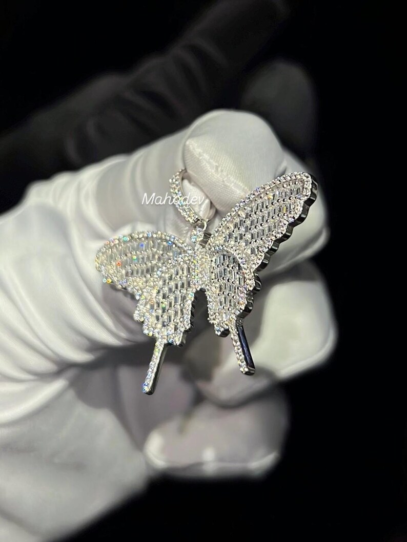May include: A sparkling butterfly pendant, encrusted with numerous clear gemstones. The pendant is held against a dark background, showcasing its intricate design and reflective qualities. The butterfly's wings and body are detailed with a dazzling array of stones.