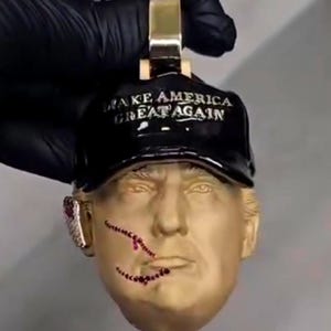 May include: A gold-toned pendant featuring a sculpted face wearing a black cap with the text "MAKE AMERICA GREAT AGAIN." The face has red gemstone accents. The pendant is held by a gold bail.
